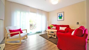 A living room with a wooden floor, a red sofa, a white armchair, and a small table with a vase. - Ferienhaus, Fedderwardersiel (Butjadingen)