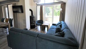 A modern living room with a grey sofa, a dining table with chairs, a black fireplace, and a large window with curtains. - Exklusives Skandinavisches Ferienhaus (Mirow)