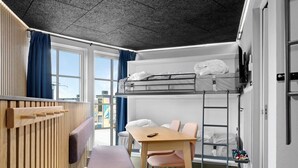 A dormitory room with bunk beds, a desk, chairs, a wooden panel wall, and a window with curtains.