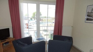Ein Zimmer mit zwei dunkelblauen Stühlen, einem Holz-TV-Möbel mit Flachbildfernseher, einem kleinen Tisch mit Laptop und einem Fenster mit roten Vorhängen.