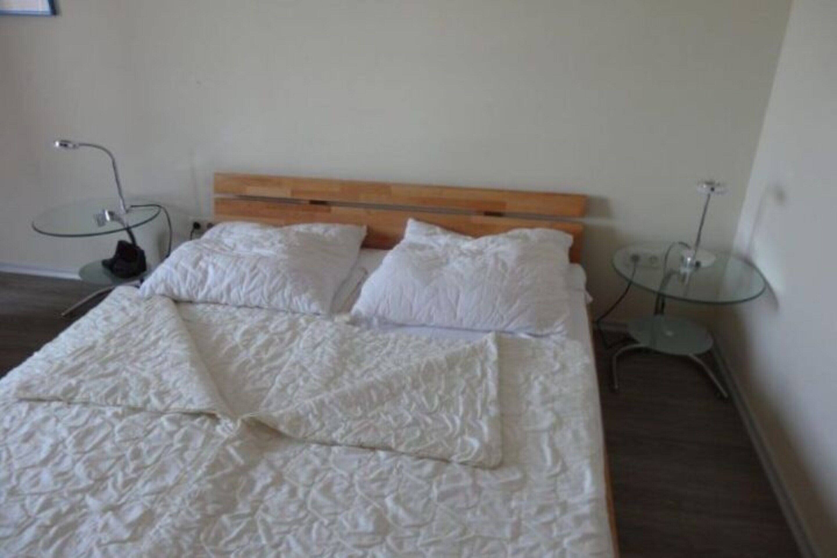 Una cama con cabecera de madera, dos almohadas blancas y una manta texturizada de color blanco. Hay dos mesitas de noche de cristal con lámparas.