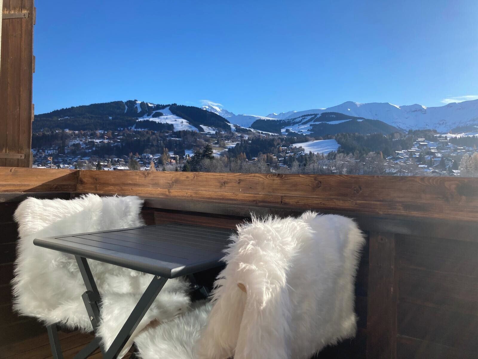 Una terraza de madera con una mesa y sillas, con vistas a un paisaje montañoso nevado y un pueblo en la falda.