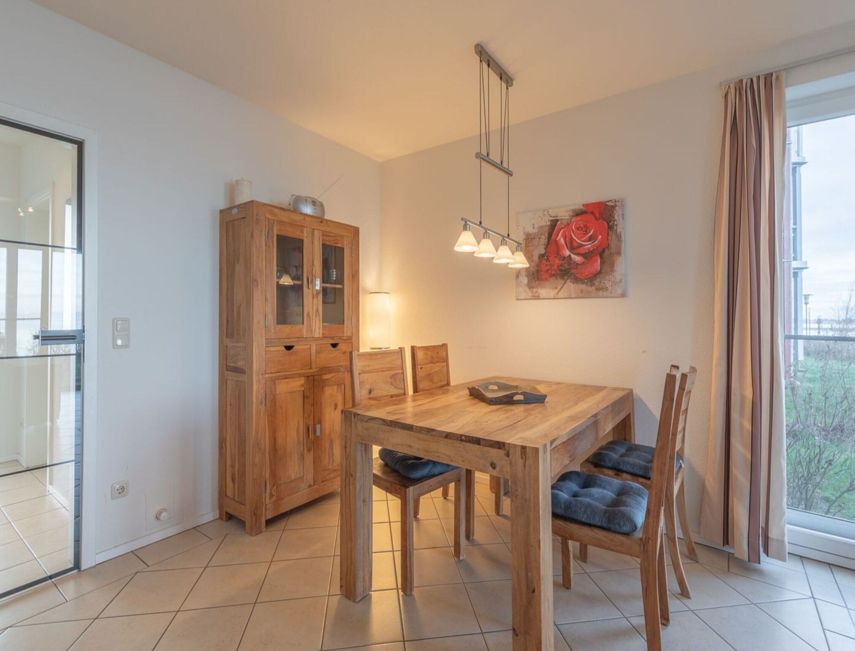 A dining area with a wooden table and chairs, a cabinet with glass doors, a hanging light fixture, and a painting of a rose on the wall.