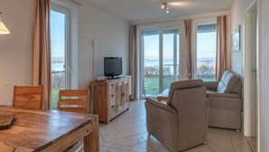 A living room with a wooden table, a sofa, a television on a wooden cabinet, and large windows with curtains. - Ferienwohnung im Ferienpark Mueritz (Rechlin)