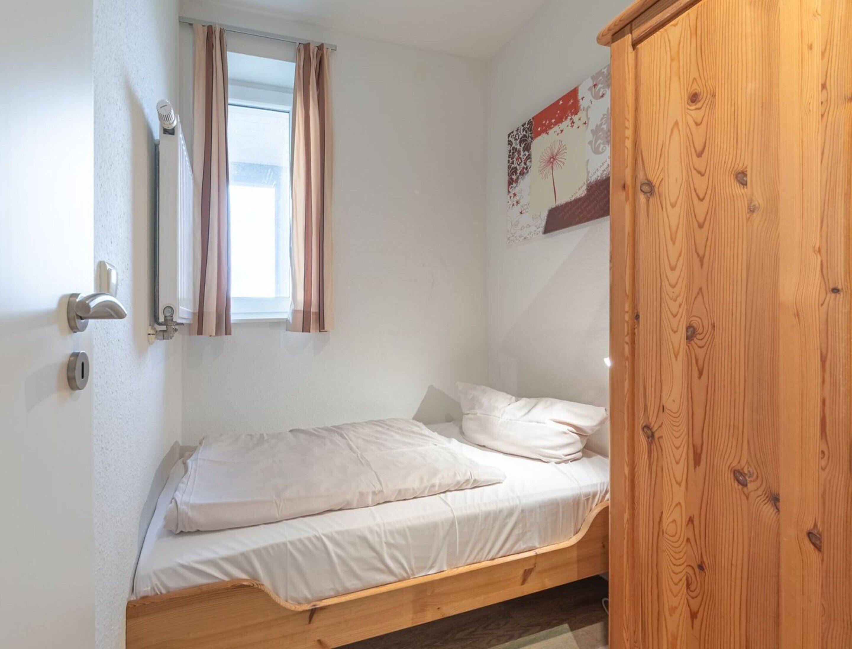 Ein kleines Zimmer mit einem Einzelbett, einem Holzbettgestell, einem Fenster mit Vorhängen, einem Heizkörper und einem Holzschrank.