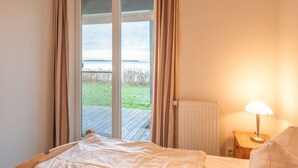 A bedroom with a bed, a nightstand with a lamp, and a view of a grassy area through a sliding glass door. - Ferienwohnung im Ferienpark Mueritz (Rechlin)