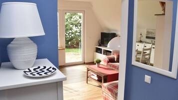 A room with a white lamp on a table, a television on a stand, and a view of a garden through a door.
