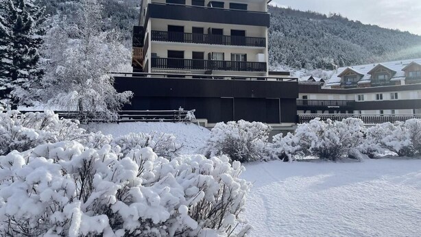 Een modern meerlaags gebouw met balkons, omgeven door met sneeuw bedekte bomen en een bergachtige achtergrond.