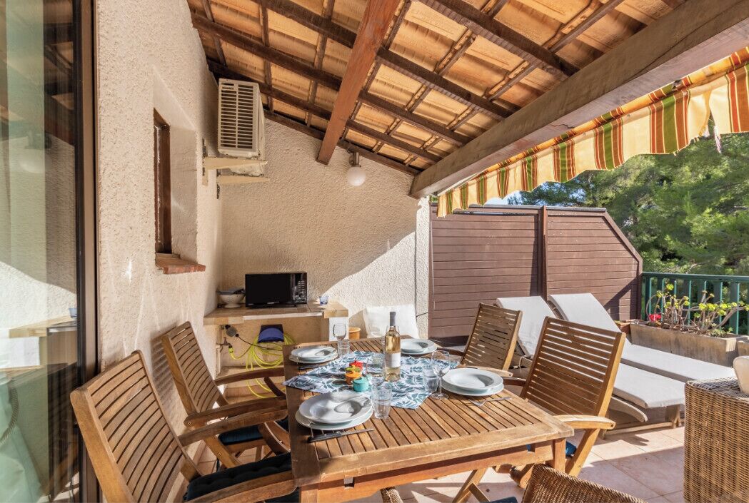 Een overdekt terras met een eettafel voor vier personen, houten stoelen en een kleine keukenhoek met een magnetron.