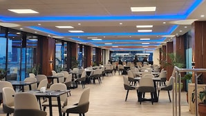 Een ruime eethoek met meerdere tafels en stoelen, een bar op de achtergrond en grote ramen met uitzicht op de stadsrand.