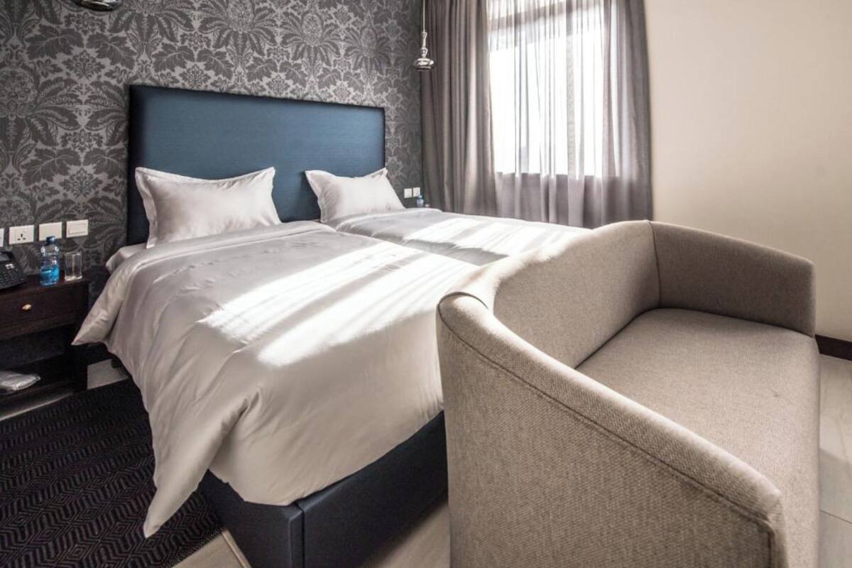 A hotel room with a double bed, a nightstand with a lamp, a chair, and a patterned wallpaper.