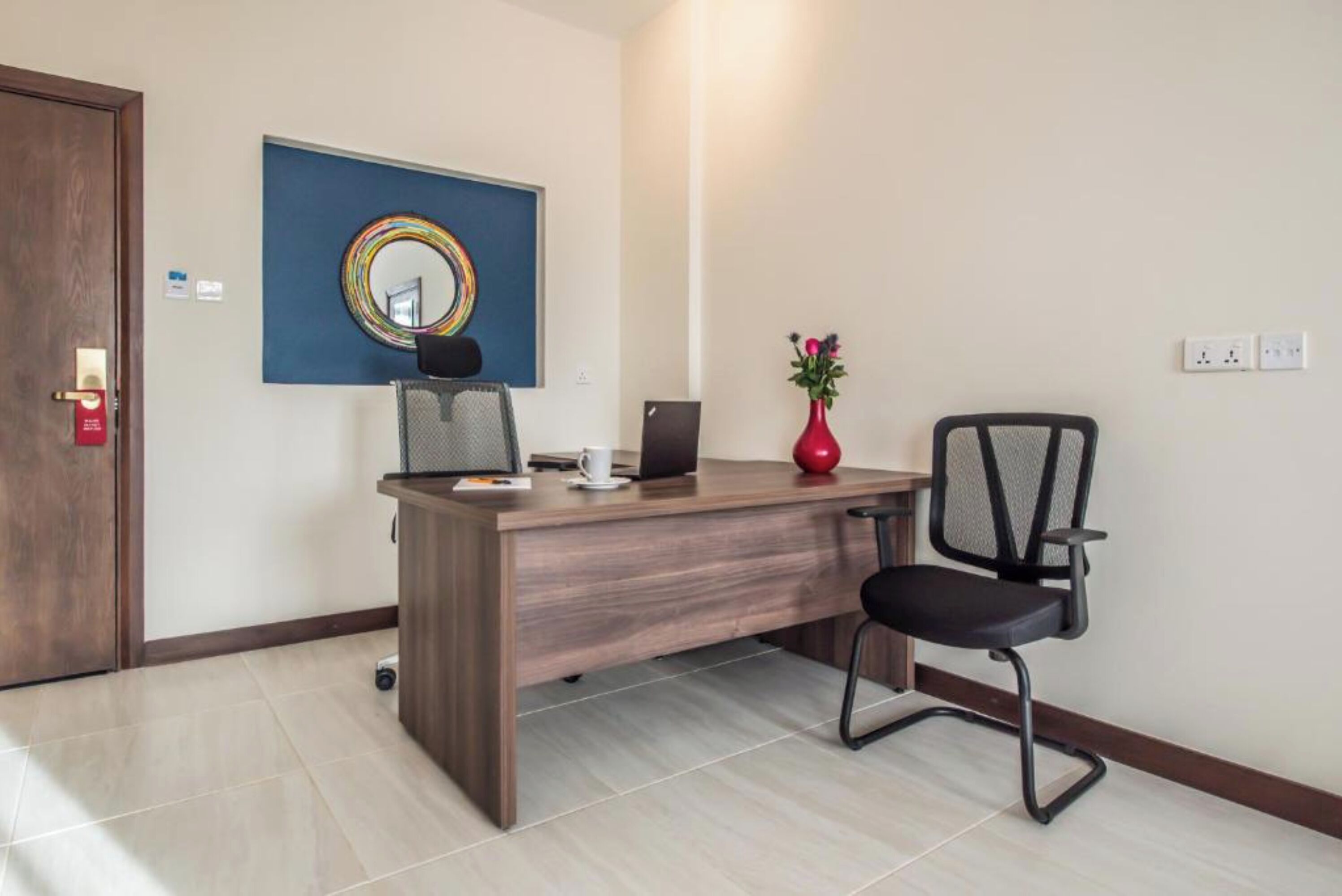 Un espacio de oficina moderno con un escritorio de madera, una silla de oficina negra, una computadora portátil, una taza de café, un jarrón con flores y un espejo montado en la pared.
