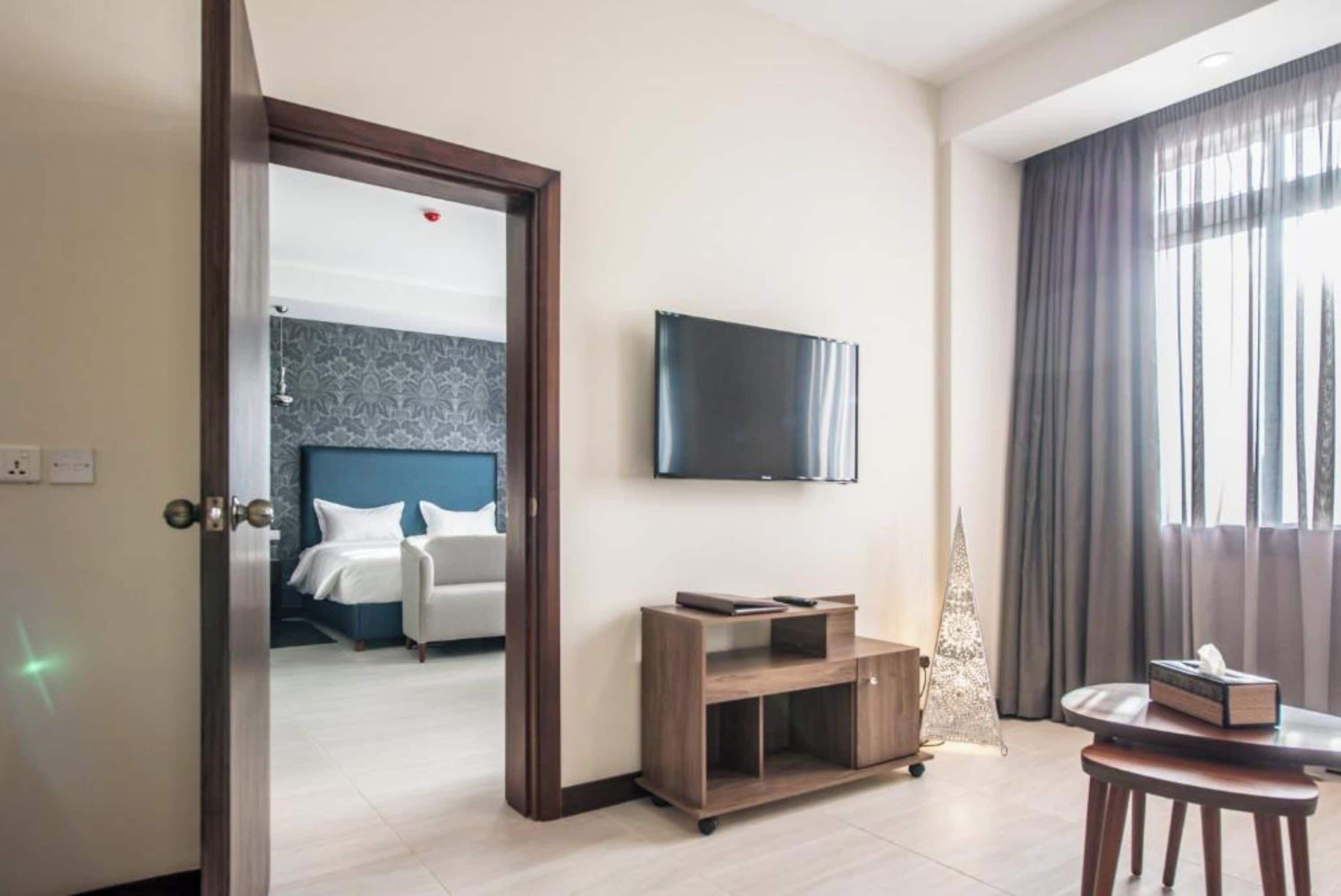 A hotel room with a bed, a TV mounted on the wall, a small wooden table, and a window with curtains.