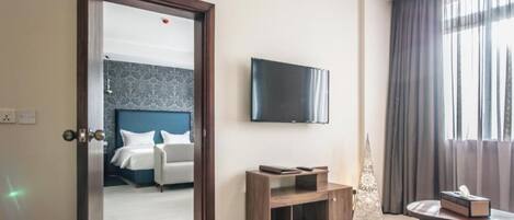 Una habitación de hotel con cama, televisor montado en la pared, una pequeña mesa de madera y ventana con cortinas.