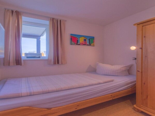 Ein Einzelbett mit Holzrahmen, ein Fenster mit Vorhängen, ein farbenfrohes Bild an der Wand und ein Holzschrank.
