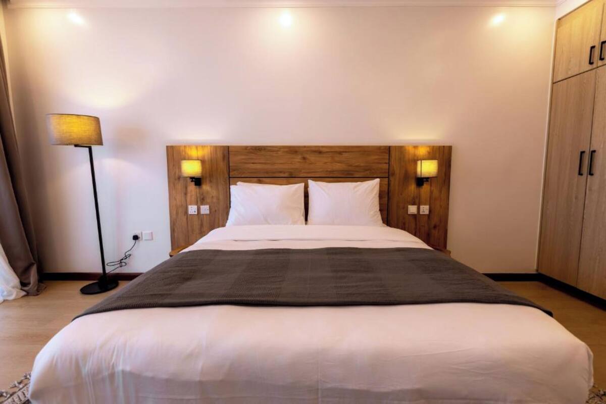 A neatly made bed with white linens and a dark throw blanket, flanked by two wall-mounted lamps and a wooden headboard.
