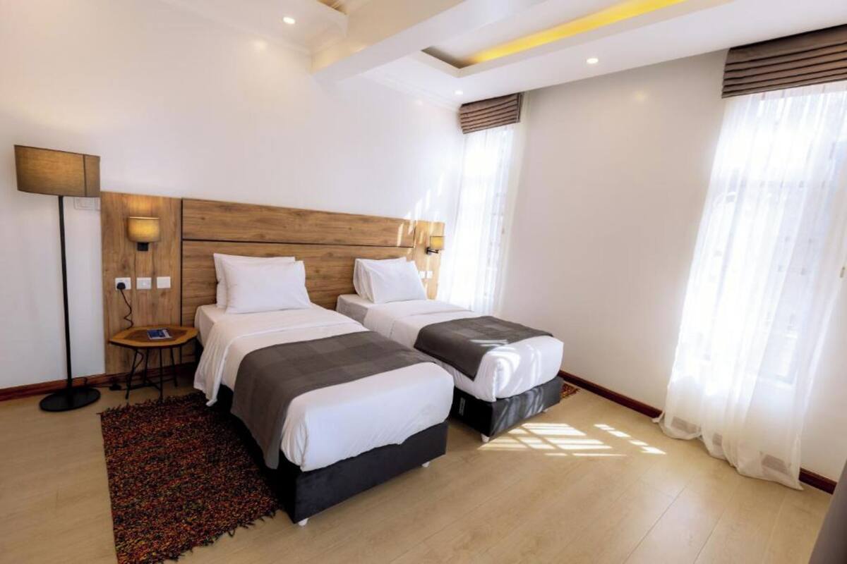 A modern hotel room with two beds, a small table, a floor lamp, and a large window with sheer curtains.