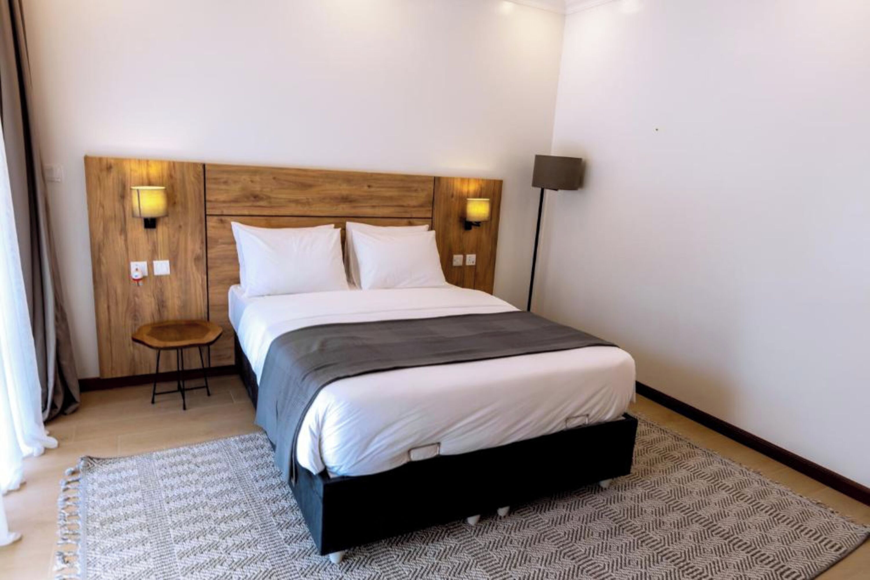 A hotel room with a wooden headboard, two bedside lamps, a small stool, and a patterned rug.