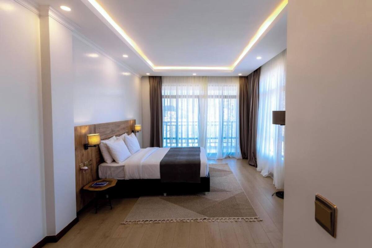 A modern hotel room with a large bed, bedside lamps, a wooden headboard, and a view of the outdoors through large windows.