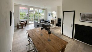 Un salon spacieux avec une table à manger en bois, une cheminée et de grandes fenêtres donnant sur un paysage verdoyant.