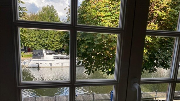 Une vue à travers une fenêtre à carreaux, donnant sur une étendue d’eau où l’on aperçoit des bateaux et un quai.
