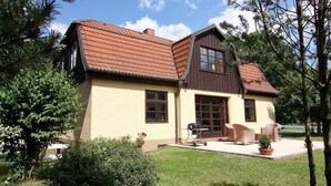 Ein zweistöckiges Haus mit einem braunen Dach, beige farbenen Wänden und einer Terrasse mit Gartenmöbeln.