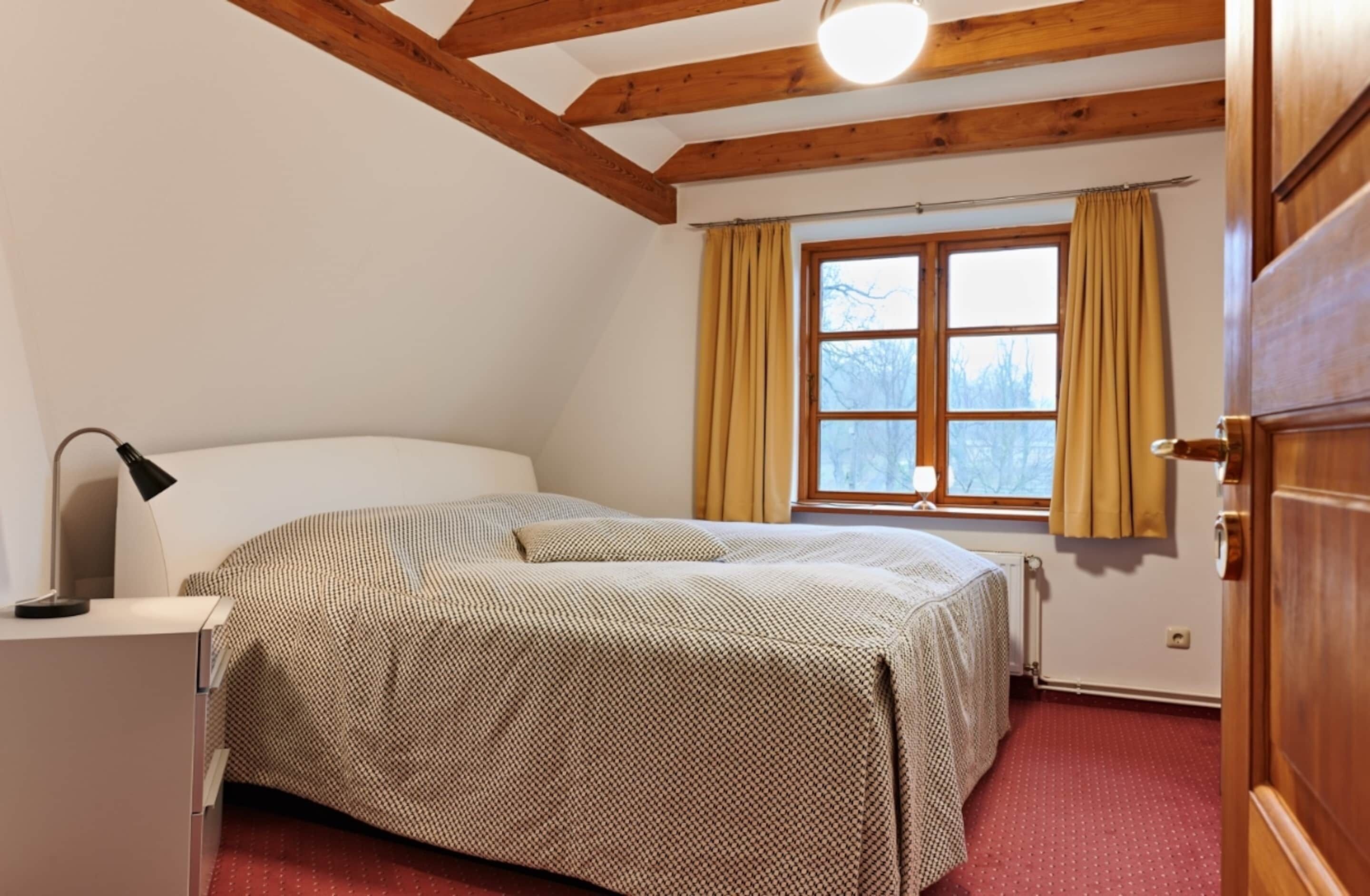 A bedroom with a bed, a desk with a lamp, a window with curtains, and wooden beams on the ceiling.