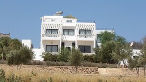 A modern two-story building with balconies, a stone wall in the foreground, and surrounding vegetation. - Air-conditioned villa with private pool 15 minutes from the beaches (Essaouira)