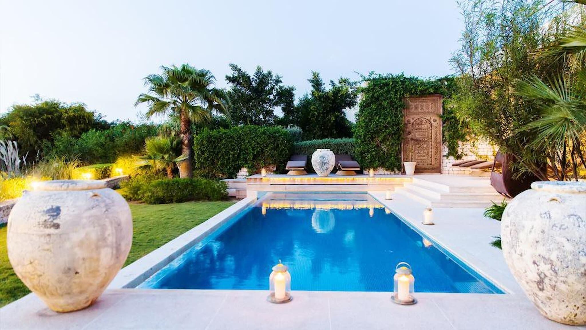 A swimming pool with two large stone vases, a seating area with sun loungers, and a well-maintained garden with various plants and a decorative wall.