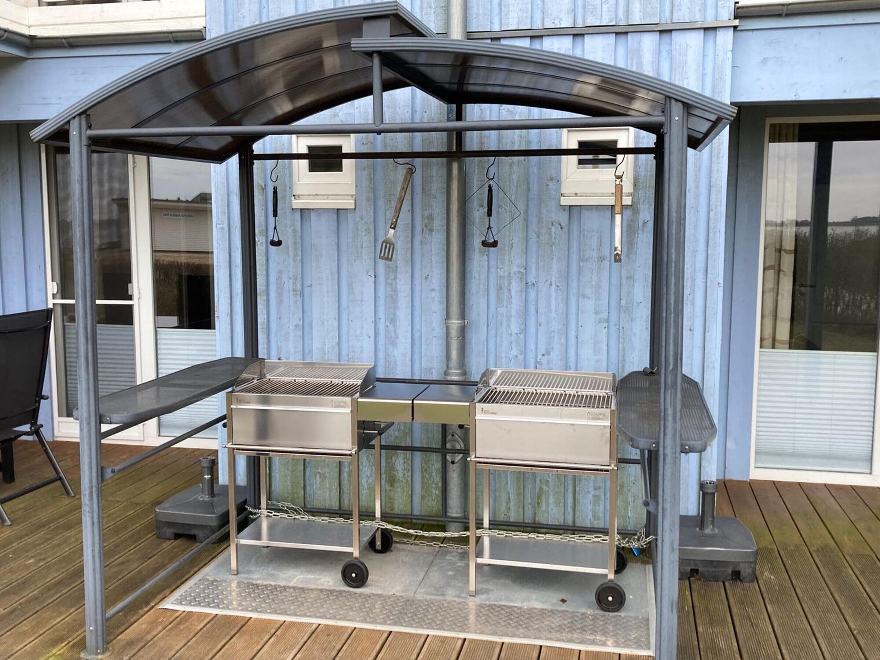 Ein überdachter Grillbereich im Freien mit Metallkonstruktion, zwei Grillstationen und einer Kettenabsperrung.