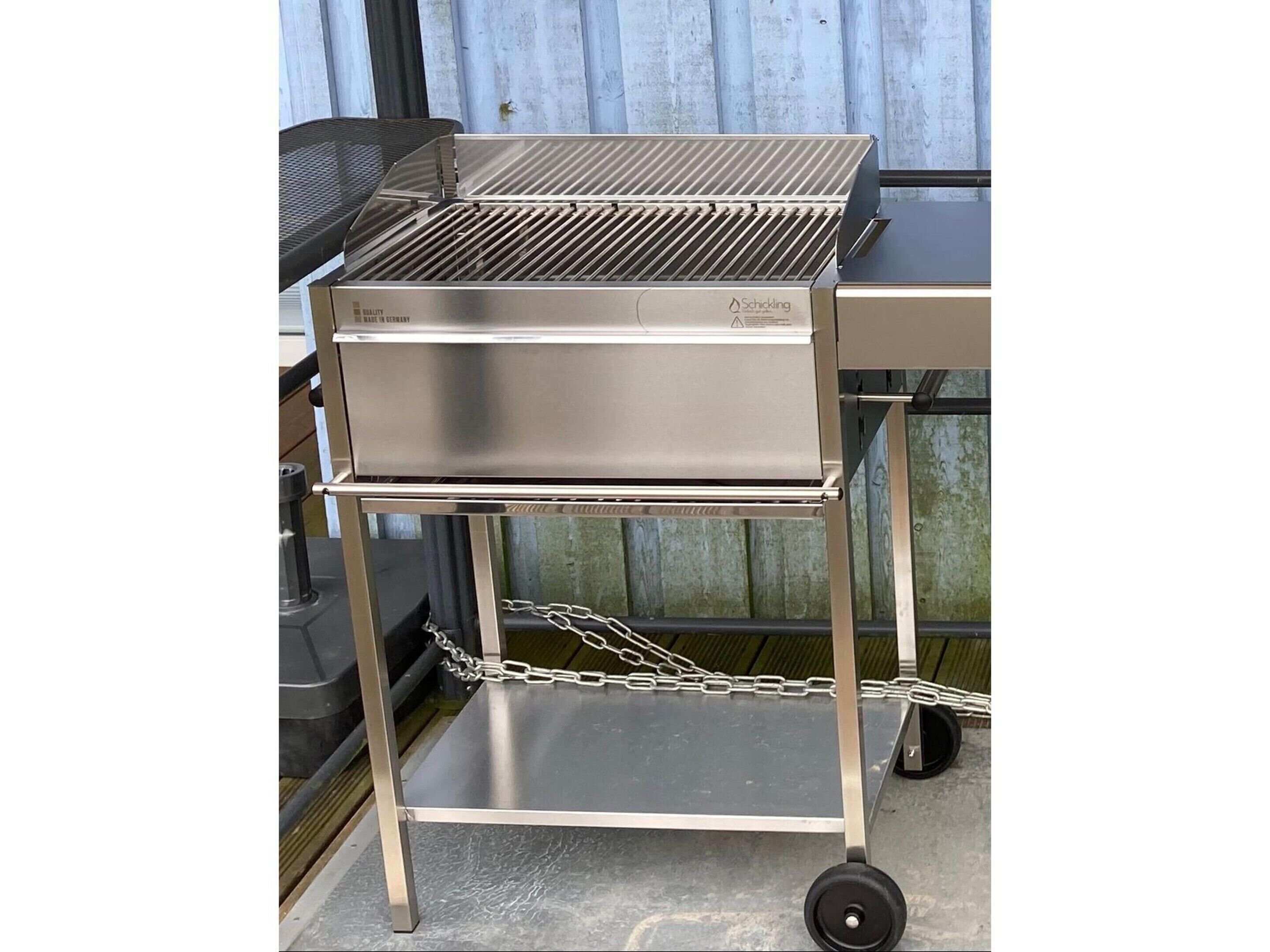 Un chariot en acier inoxydable pour barbecue, doté d’une surface de cuisson plane et d’une étagère inférieure, fixé à l’aide de chaînes, sur un fond en métal ondulé.