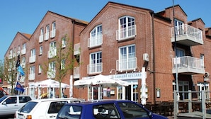 A brick building with multiple windows and balconies, a storefront with outdoor seating, and parked cars. - Ferienwohnung in Orth for Zwei Personen (Fehmarn)