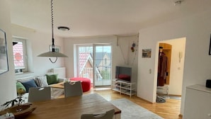 A modern living room with a wooden dining table, a red bean bag, a TV, and a large window with a view of a house. - Ferienwohnung in Orth for Zwei Personen (Fehmarn)