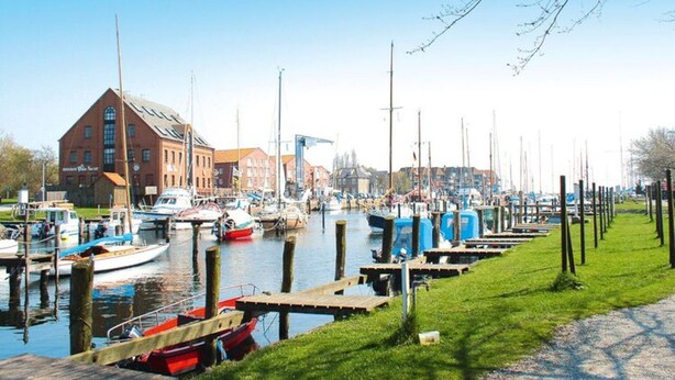 A marina with boats docked, a building with a red facade, and a grassy area with wooden posts. - Ferienwohnung in Orth for Zwei Personen (Fehmarn)