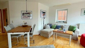 A living room with a wooden dining table, white sofa, and a red bean bag. - Ferienwohnung in Orth for Zwei Personen (Fehmarn)