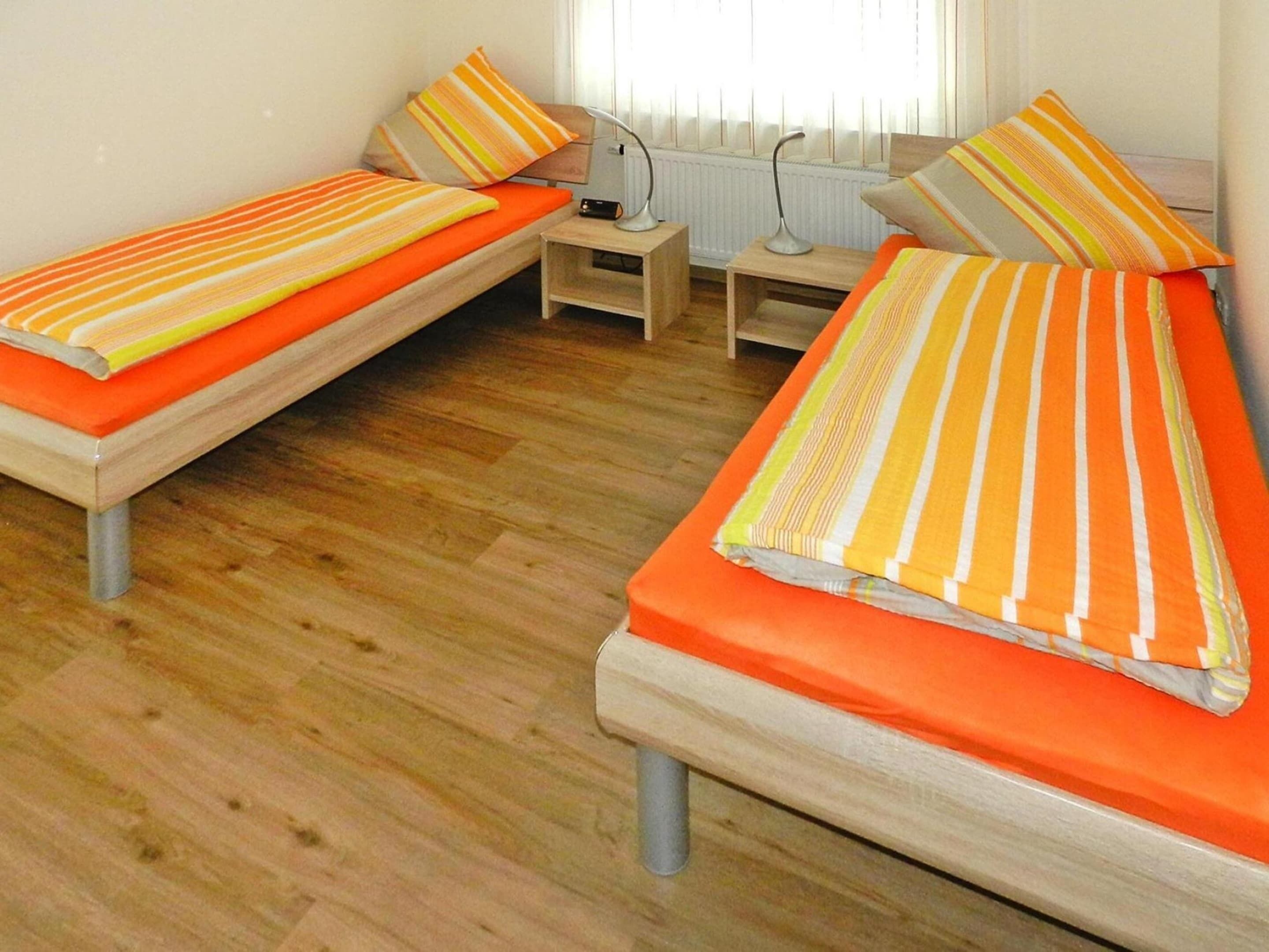 Une chambre avec deux lits simples, chacun agrémenté de draps et d’oreillers à rayures orange. Une petite table de chevet en bois avec une lampe se trouve entre les deux lits.