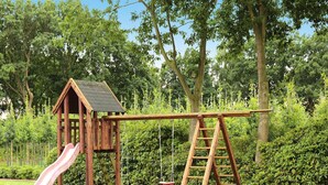 A wooden play structure with a slide and a swing set in a grassy area with trees in the background. - Ferienwohnung Landblick, Rastede (Rastede)