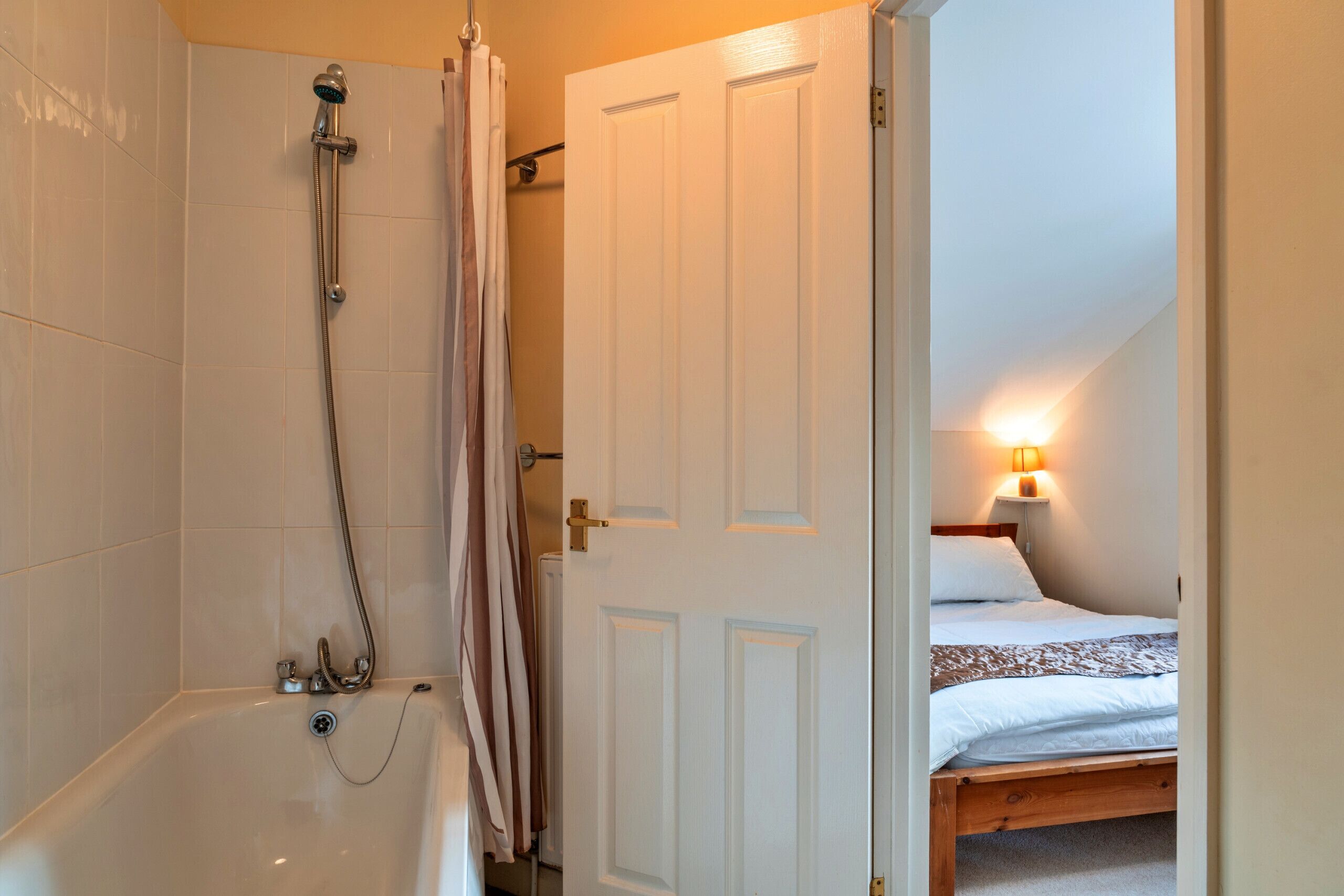 A bathroom with a bathtub, shower, and a door leading to a bedroom with a bed and a nightstand.