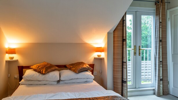 A bedroom with a bed, two bedside lamps, and a view of the outdoors through a door. - Forest Park Lodge (Umberleigh)