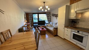 Een gezellige woonkamer met een houten eettafel, een bank en een televisie. Er is een keuken met witte kastjes en een fornuis te zien.