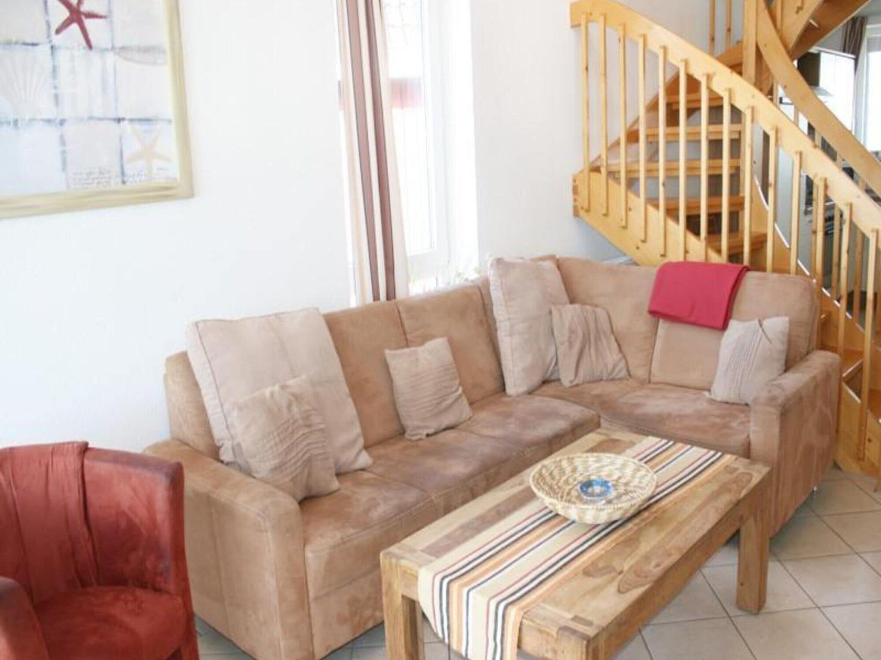 A living room with a beige sofa, a wooden coffee table, and a red chair. A wooden staircase is visible in the background.