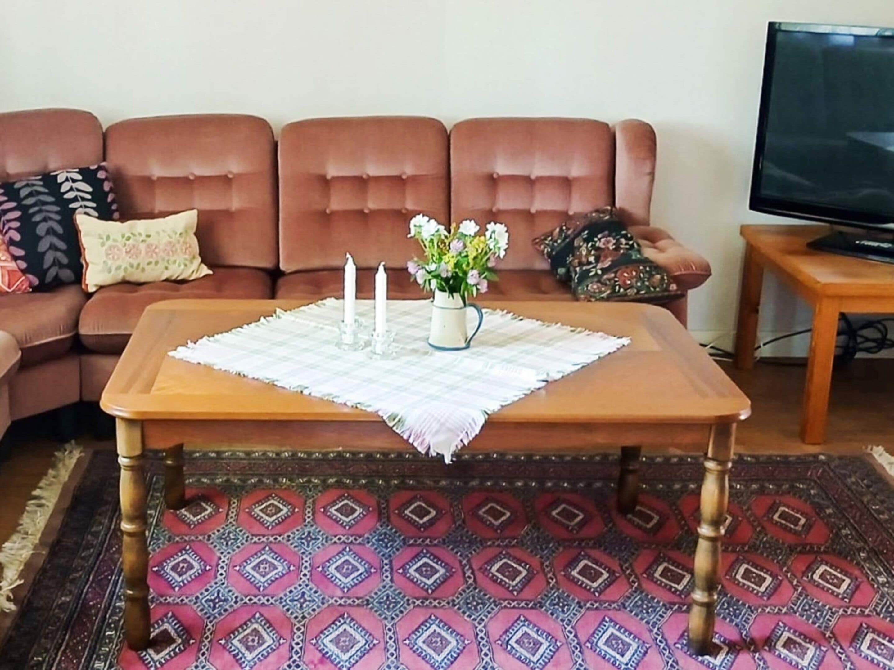 A living room with a brown sofa, a wooden coffee table with a placemat, a vase with flowers, and a television on a wooden stand.