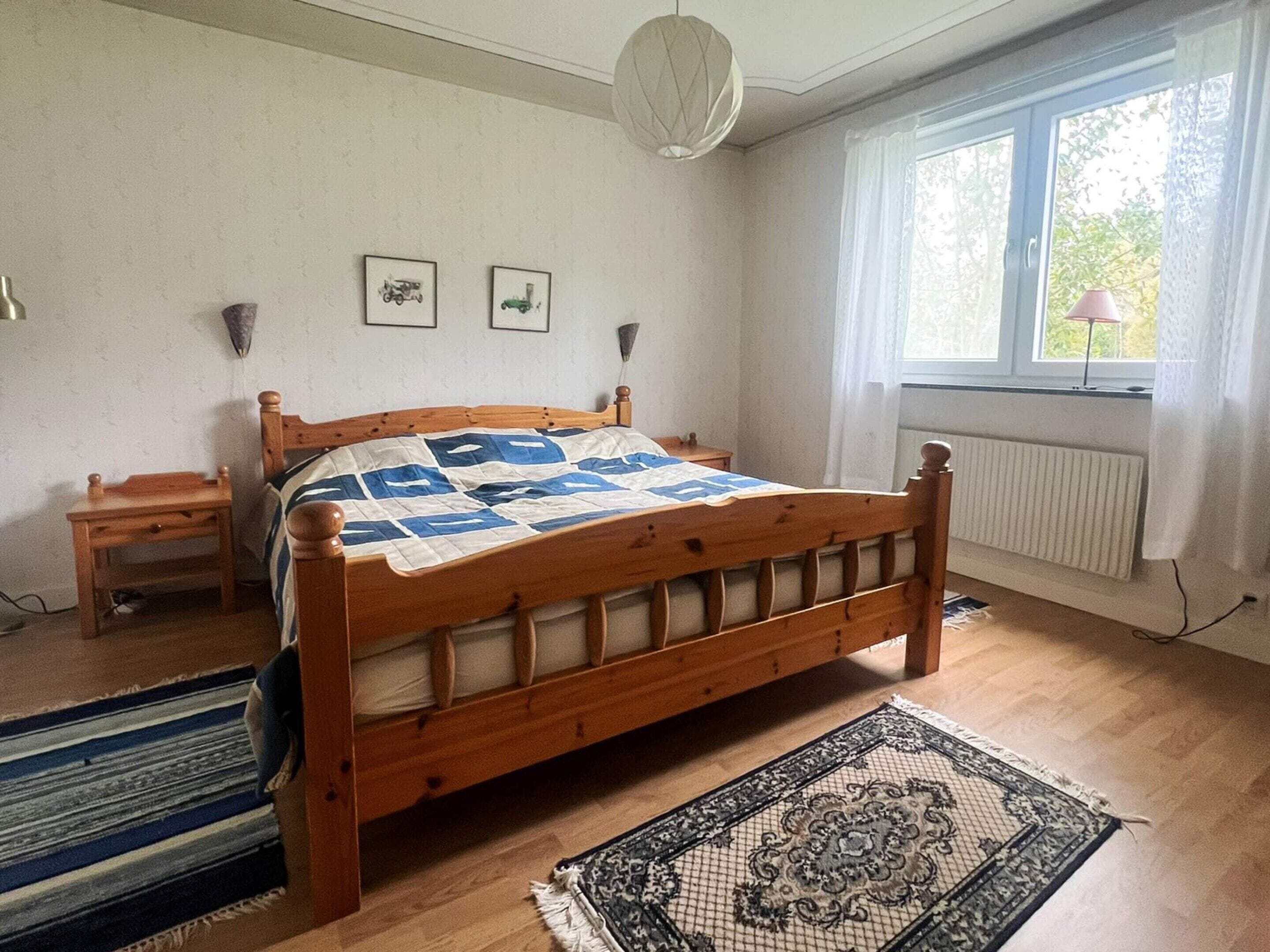 A bedroom with a wooden bed, a bedside table, a nightstand, a window with curtains, and a radiator.