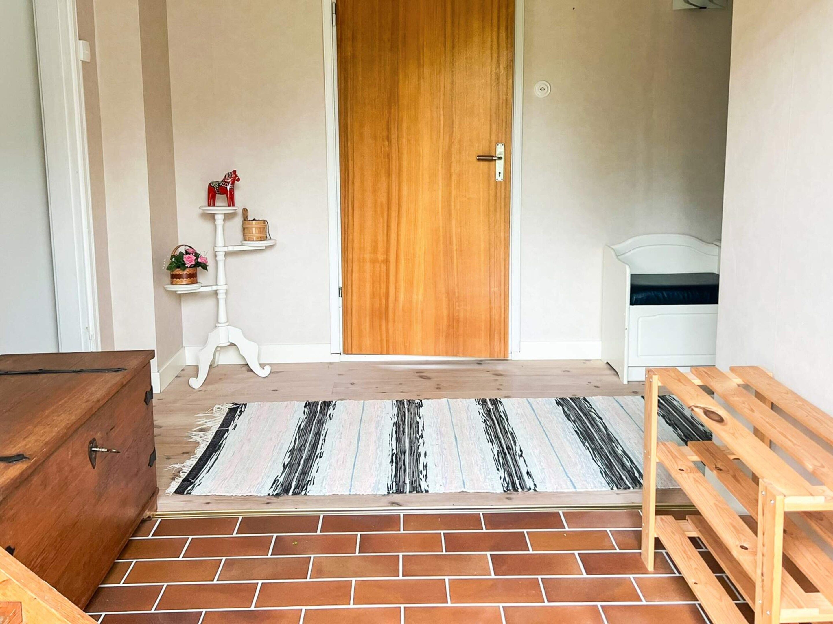A room with a wooden door, a white bedside table, a small shelf with decorative items, and a patterned rug on the floor.