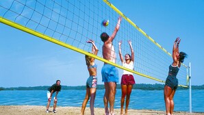 Un grupo de personas jugando voleibol de playa en una arena dorada, con un cielo azul despejado y agua tranquila al fondo.