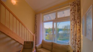 Una habitación con escalera de madera, un sofá color beige y un ventanal con vistas a árboles y una masa de agua.