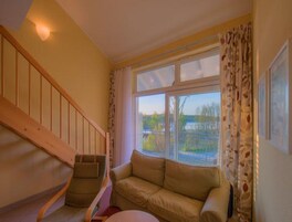 A room with a wooden staircase, a beige sofa, and a large window with a view of trees and a body of water.