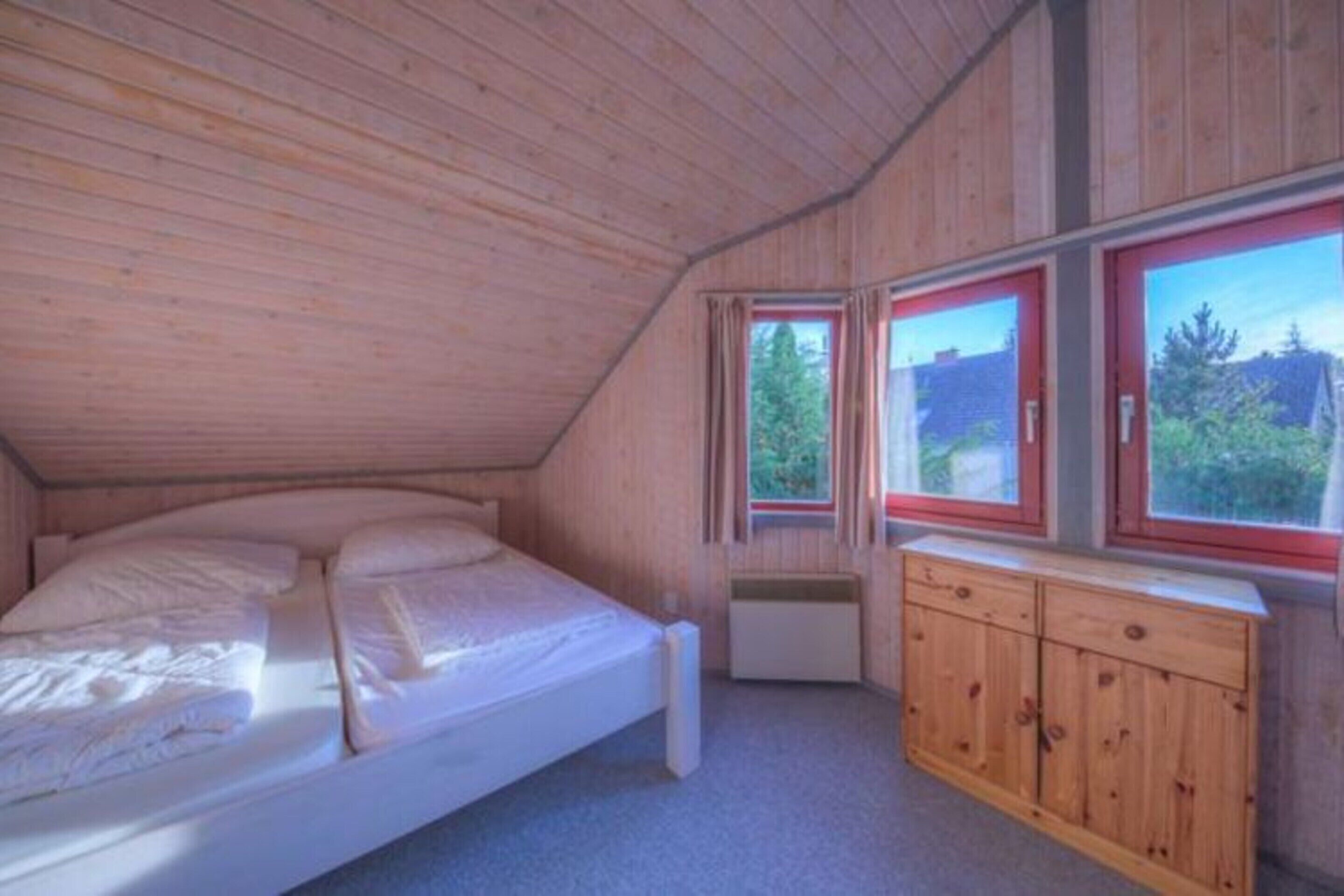 A bedroom with a wooden ceiling, a bed, a dresser, and windows with red frames.
