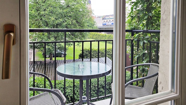 A balcony with a glass table and wicker chairs, overlooking a green lawn and a building with a tower. - DReAM Post Aparthotel (Kraków)