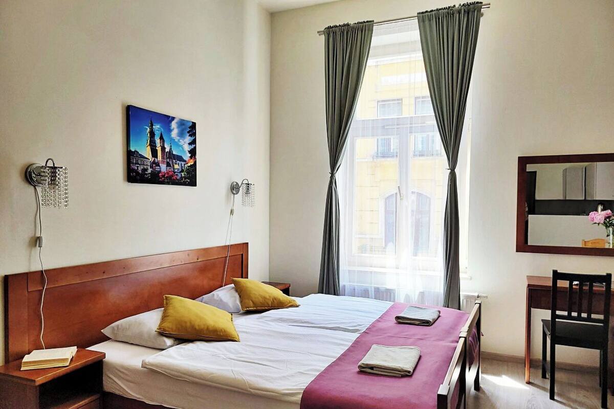 Camera d'albergo con un letto grande, due cuscini, un comodino con un libro, una scrivania con una sedia, una finestra con tende e un quadro appeso al muro.